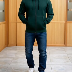 May include: A person wearing a dark green zip-up hoodie with a front pocket, a navy blue t-shirt, and blue jeans. The person is also wearing white sneakers. The background is a light brown wooden wall.