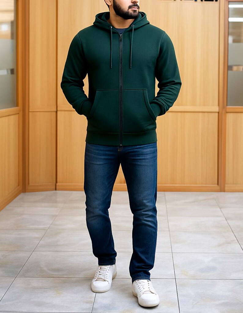May include: A person wearing a dark green zip-up hoodie with a front pocket, a navy blue t-shirt, and blue jeans. The person is also wearing white sneakers. The background is a light brown wooden wall.