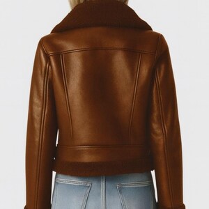 May include: A brown faux leather jacket with a shearling collar and trim. The jacket has a classic biker style with a zip closure and a tailored fit. The model is wearing light blue jeans.