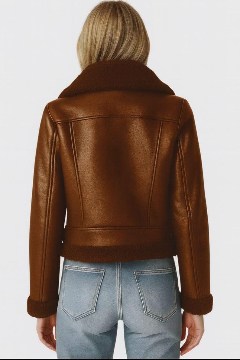 May include: A brown faux leather jacket with a shearling collar and trim. The jacket has a classic biker style with a zip closure and a tailored fit. The model is wearing light blue jeans.