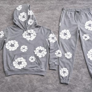 May include: A gray hoodie and matching sweatpants set. The hoodie has a drawstring hood and a front pocket. Both pieces are decorated with a white floral pattern. The sweatpants have a drawstring waist and cuffed ankles. The set is made of a soft material.