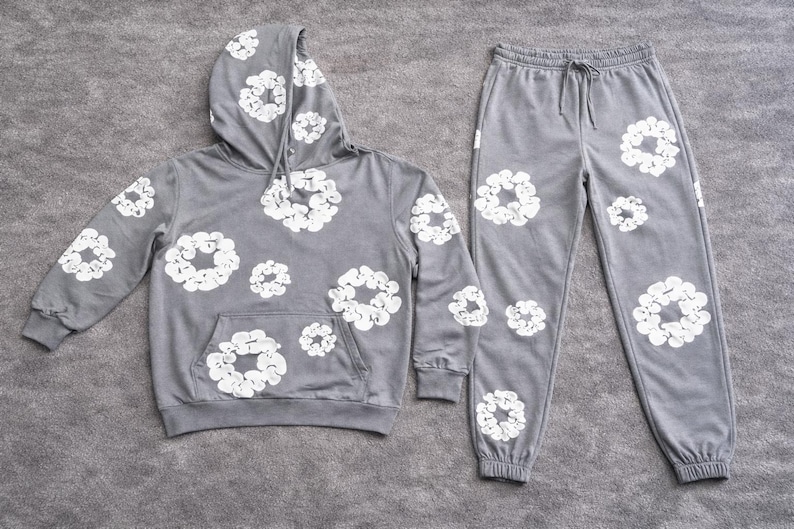 May include: A gray hoodie and matching sweatpants set. The hoodie has a drawstring hood and a front pocket. Both pieces are decorated with a white floral pattern. The sweatpants have a drawstring waist and cuffed ankles. The set is made of a soft material.