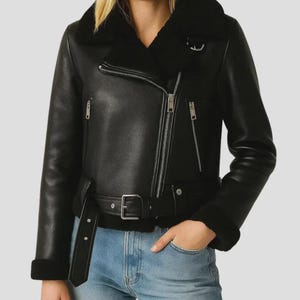 May include: Black leather jacket with a shearling collar and lining. The jacket features a diagonal zipper closure, a belt at the waist, and zippered pockets. The sleeves have shearling cuffs. The model is wearing blue jeans.