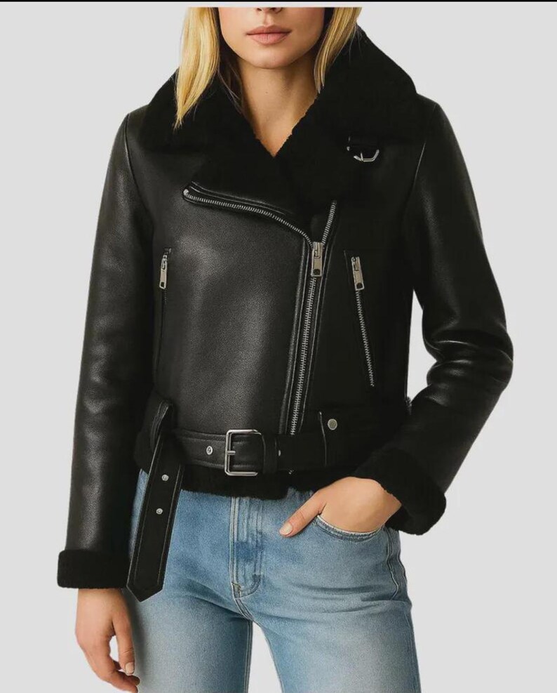 May include: Black leather jacket with a shearling collar and lining. The jacket features a diagonal zipper closure, a belt at the waist, and zippered pockets. The sleeves have shearling cuffs. The model is wearing blue jeans.