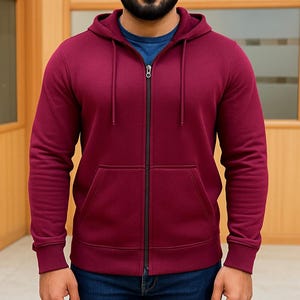 May include: A burgundy zip-up hoodie with a drawstring hood and front pockets. The hoodie has a black zipper and is worn over a blue crew neck shirt. The person is wearing blue jeans.
