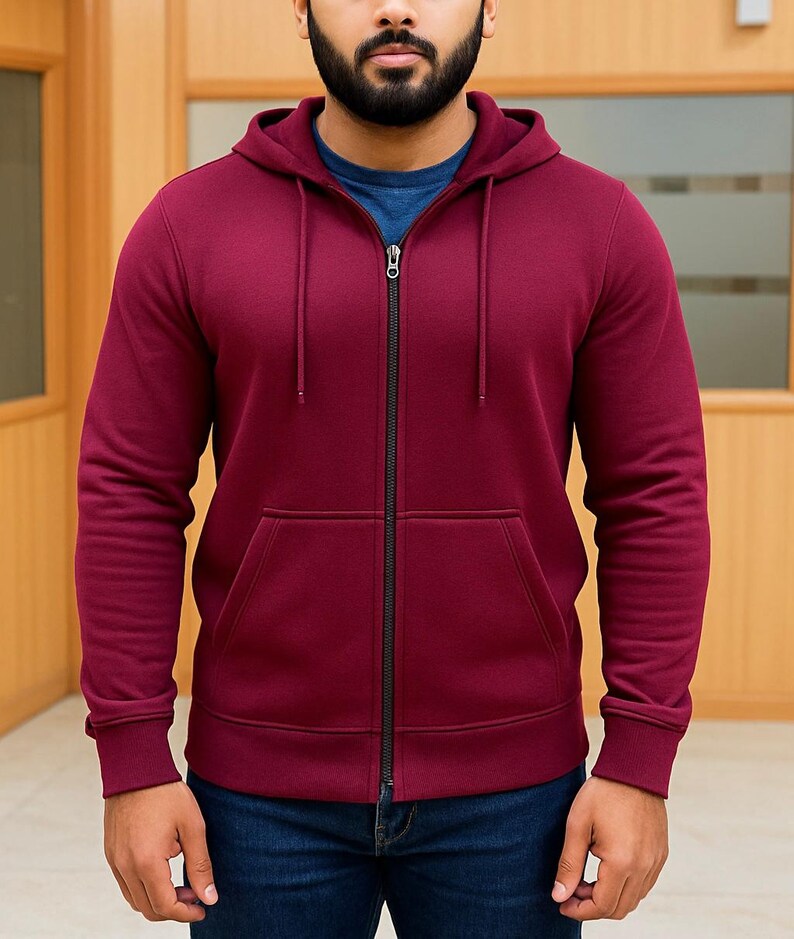 May include: A burgundy zip-up hoodie with a drawstring hood and front pockets. The hoodie has a black zipper and is worn over a blue crew neck shirt. The person is wearing blue jeans.