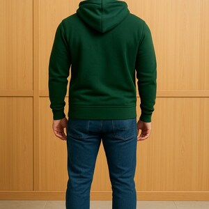 May include: A person wearing a dark green hooded sweatshirt and blue jeans. The sweatshirt has a drawstring hood and ribbed cuffs and hem. The jeans are a classic five-pocket style. The person is standing in front of a light brown wooden wall.