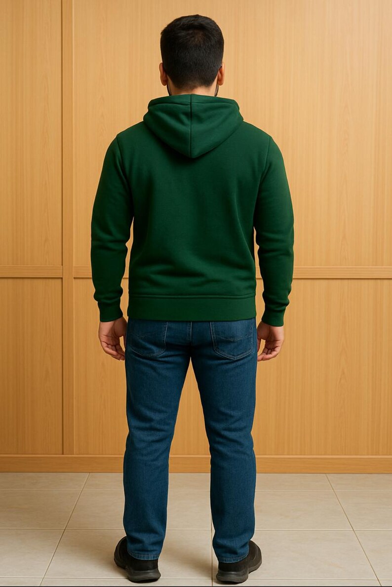 May include: A person wearing a dark green hooded sweatshirt and blue jeans. The sweatshirt has a drawstring hood and ribbed cuffs and hem. The jeans are a classic five-pocket style. The person is standing in front of a light brown wooden wall.