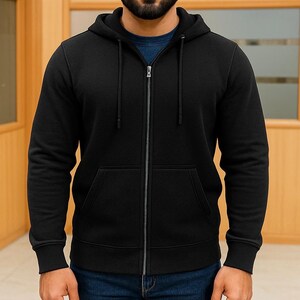 May include: A black zip-up hoodie with a drawstring hood and front pockets. The hoodie is made of a soft material and has a full-length zipper. The sleeves have ribbed cuffs. The person is wearing blue jeans and a blue shirt underneath.