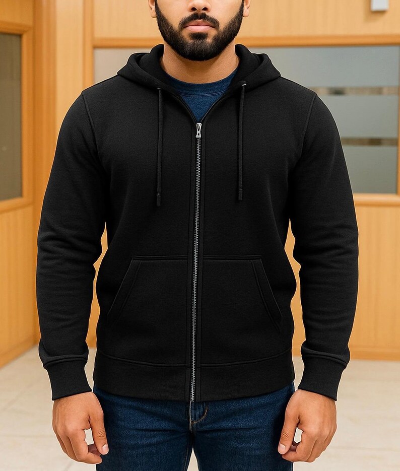 May include: A black zip-up hoodie with a drawstring hood and front pockets. The hoodie is made of a soft material and has a full-length zipper. The sleeves have ribbed cuffs. The person is wearing blue jeans and a blue shirt underneath.