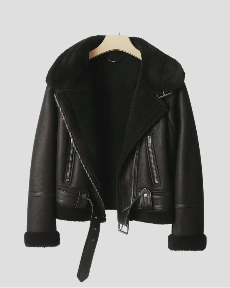 May include: Black faux leather jacket with a shearling collar and lining. The jacket features a zip closure, a belt at the waist, and zip pockets. The cuffs are trimmed with shearling. The jacket is hanging on a wooden hanger.