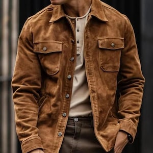 May include: A brown suede jacket with two chest pockets and button closures. The jacket is worn over a light-colored shirt. The jacket has a classic collar and a tailored fit, suitable for casual or semi-formal wear.