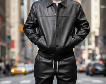 Può includere: Un completo in pelle nera composto da una giacca abbottonata con colletto e tasche, e pantaloni abbinati con coulisse in vita. L'outfit è completato da scarpe da ginnastica bianche. La persona è in piedi su una strada cittadina.