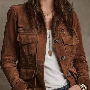 May include: A brown suede jacket with multiple pockets and button fastenings. The jacket has a tailored fit and is worn over a white top and blue jeans. A gold necklace and bracelets add a touch of elegance to the outfit.