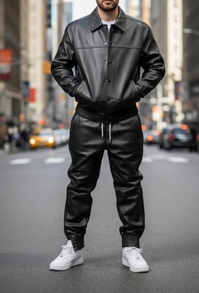 May include: A black leather jacket with a button-up front and matching black leather pants. The jacket has a collar and pockets. The pants have a drawstring waist. The outfit is paired with white sneakers. The person is standing on a city street.