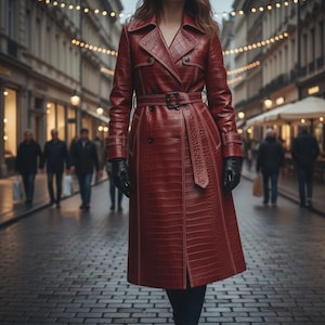 Pode incluir: Um trench coat vermelho com padrão de crocodilo, cinto e luvas pretas. O casaco tem um design clássico com frente de abotoamento duplo e gola entalhada. O casaco é comprido até ao joelho e parece ser de couro.