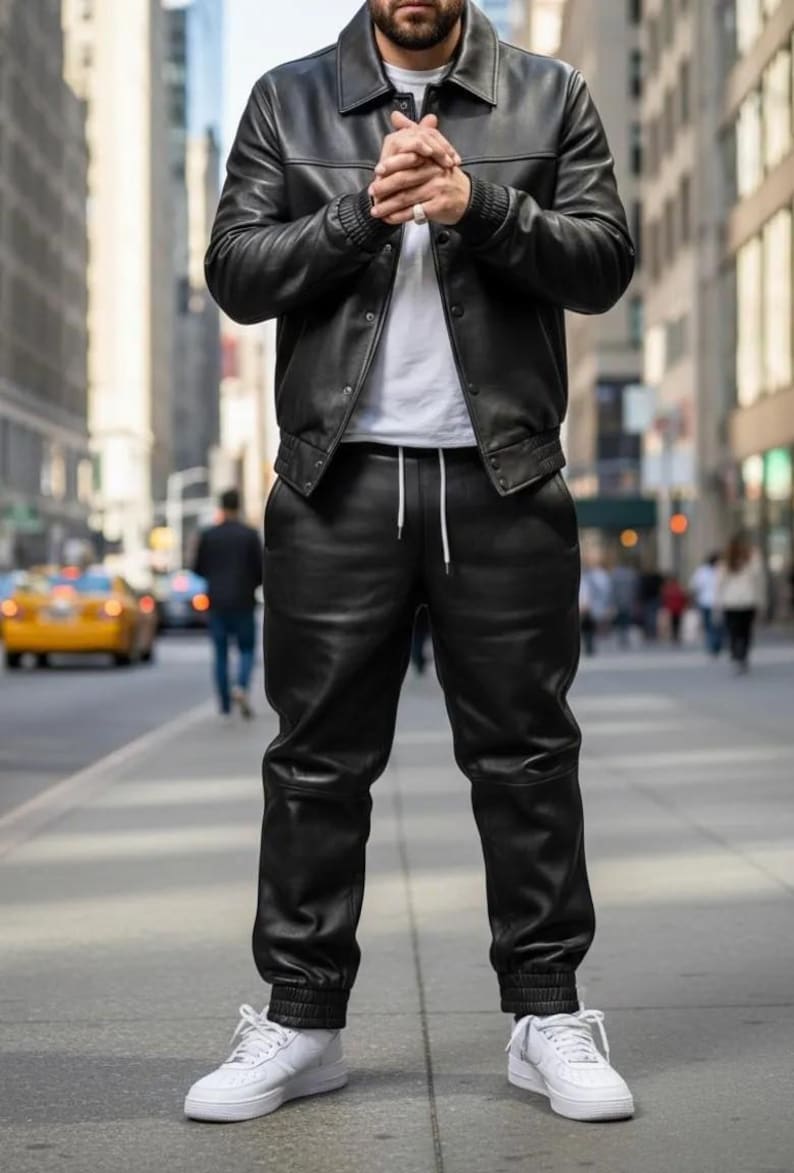 May include: A person wearing a black leather jacket and matching leather pants with white sneakers. The jacket has a collar and button closure. The pants have a drawstring waist and elastic cuffs. The background is a city street.