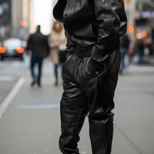 May include: A person wearing a black leather jacket, matching pants, and a black cap. The outfit is paired with white sneakers. The person is standing on a city street.