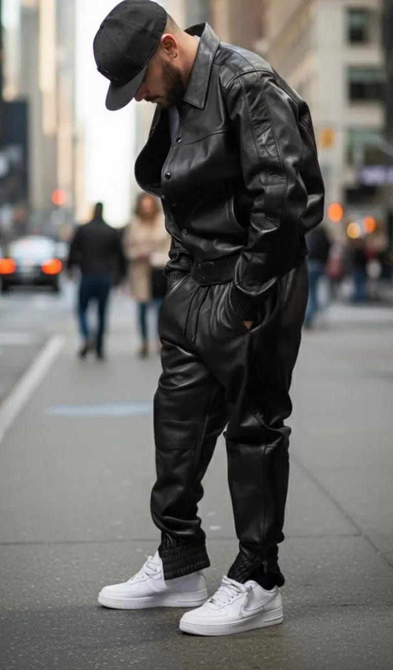 May include: A person wearing a black leather jacket, matching pants, and a black cap. The outfit is paired with white sneakers. The person is standing on a city street.