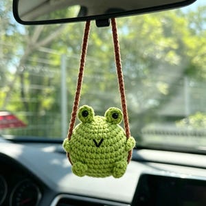 May include: A round, light green crocheted frog ornament hanging from a brown yarn string. The frog features two small eyes with black pupils, a simple V-shaped black stitched mouth, and small limbs, suspended from a rearview mirror.