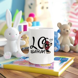 Pu&ograve; includere: Tazza in ceramica bianca con un design a scritta nera "Love Sarah" e un'illustrazione di Minnie Mouse. La tazza &egrave; circondata da giocattoli per bambini, tra cui un coniglietto bianco e un orsacchiotto.