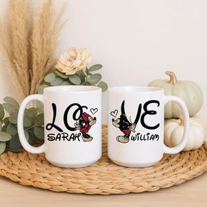 Pu&ograve; includere: Due tazze in ceramica bianca con la parola "LOVE" in corsivo nero, ognuna con un personaggio dei cartoni animati. Una tazza dice "Sarah" e l'altra "William". Le tazze sono su un vassoio intrecciato con zucche e piante secche sullo sfondo.