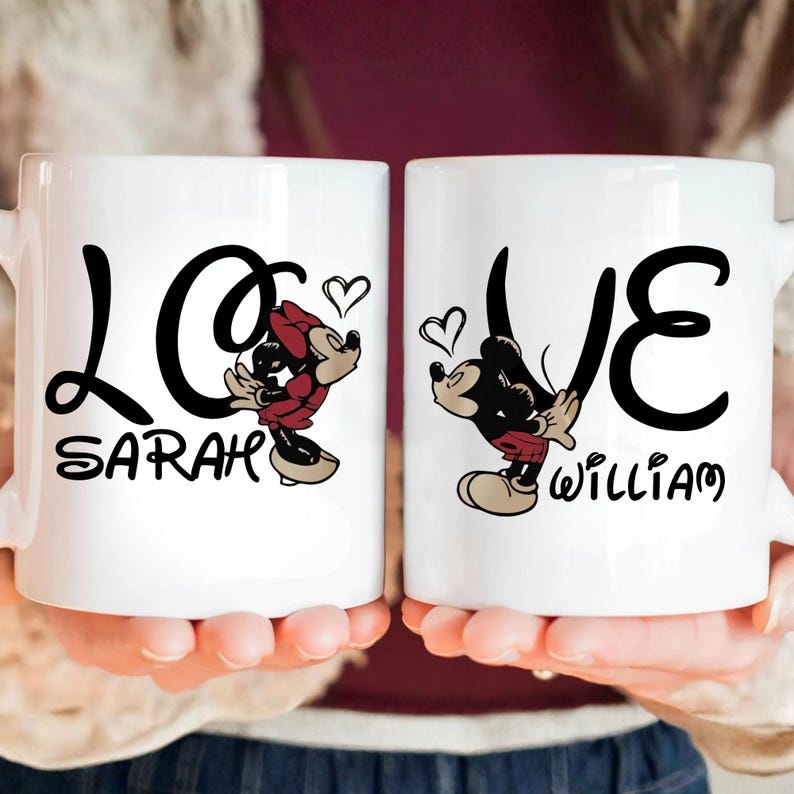 Pu&ograve; includere: Due tazze in ceramica bianca con testo nero che recita "LOVE" con i nomi "Sarah" e "William" sotto. Ogni tazza presenta un personaggio dei cartoni animati, Minnie e Topolino, con un cuore. Le tazze sono tenute da una persona.