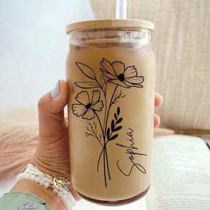 May include: A clear glass tumbler filled with a light brown beverage, featuring a black floral design and the name "Sophia" in script. The tumbler has a wooden lid and a straw. The glass is held by a person wearing a bracelet.