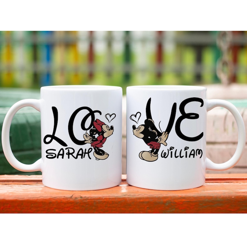 Pu&ograve; includere: Due tazze in ceramica bianca con testo nero e illustrazioni di cartoni animati. Una tazza dice "LOVE Sarah" con una grafica di Minnie Mouse, e l'altra dice "LOVE William" con una grafica di Topolino. Entrambe le tazze hanno un simbolo a forma di cuore.