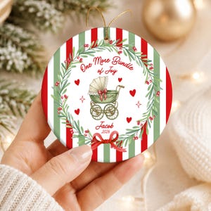 May include: A round ceramic Christmas ornament featuring red and green vertical stripes. The centre displays a green vintage baby carriage, a wreath of holly, red hearts, and the text One More Bundle of Joy, Jacob, and 2026.