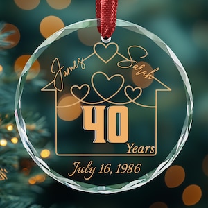 May include: A circular clear glass ornament with a faceted edge, featuring a red ribbon hanger. The etched design shows a house outline with hearts, the names James and Sarah, the number 40, the word Years, and the date July 16, 1986.