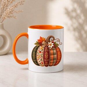 Könnte beinhalten: Ein weißer Keramikbecher mit einem leuchtend orangefarbenen Henkel und Innenrand. Die Außenseite zeigt eine stilisierte Kürbisgrafik, die mit verschiedenen herbstlichen Mustern wie Karos, Blumenmotiven und einfarbigen Abschnitten verziert ist.