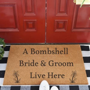 May include: A rectangular tan coir doormat featuring the black printed text A Bombshell Bride & Groom Live Here, with a simple black line drawing of a flower on the bottom left and bottom right corners of the mat surface.