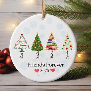 May include: A white, round Christmas ornament with four illustrated Christmas trees, each with a unique design. The ornament has the words "Friends Forever 2025" and names below each tree. A string is attached for hanging.