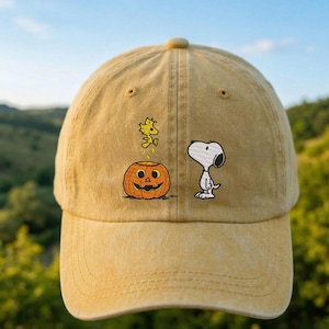 May include: A mustard yellow baseball cap featuring an embroidered design of a white beagle standing next to an orange jack-o'-lantern. A small yellow bird is depicted jumping above the pumpkin. The cap has a six-panel construction and a curved brim.