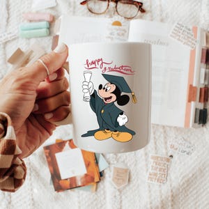 May include: White ceramic mug featuring a graduation-themed Mickey Mouse illustration. Mickey is wearing a graduation cap and gown, holding a diploma. The mug has the words "happy graduation" in red script.
