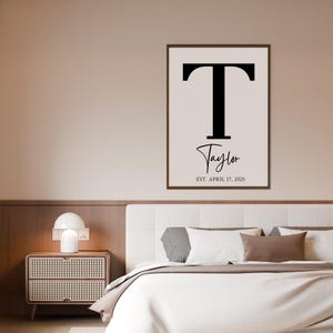 May include: A rectangular wall print in a dark brown frame featuring a large black capital letter T, the name Taylor in a black cursive script, and the text EST. APRIL 17, 2025 in a small black serif font on a light cream background.