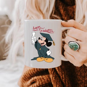 May include: White ceramic mug featuring Mickey Mouse in a graduation cap and gown, holding a diploma. The words "happy graduation" are written in red script above the character. The mug has a white handle.
