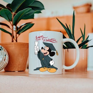 May include: White ceramic mug featuring Mickey Mouse in a graduation cap and gown, holding a diploma. The mug has the words "happy graduation" in script. The design is in full color.
