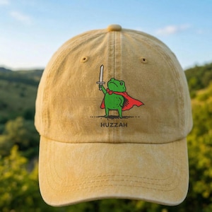 May include: A mustard yellow baseball cap featuring a graphic of a green frog wearing a red cape and holding a silver sword. Below the frog, the word HUZZAH is printed in black capital letters. The cap has a distressed, washed-out fabric texture.