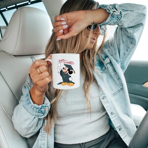 May include: White ceramic mug featuring Mickey Mouse in a graduation cap and gown, holding a diploma. The mug has the text "Happy Graduation" in red script. The mug is held by a person in a denim jacket.