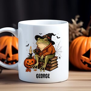 May include: A white ceramic mug featuring a graphic of a green frog wearing a black witch hat and an orange sweater, sitting on a stack of books. The frog holds a small cup, with a jack-o'-lantern, a candle, and a spiderweb nearby. The name GEORGE is printed below.