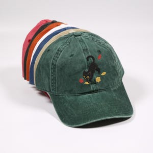 May include: A stack of baseball caps in various colors including red, black, orange, white, blue, beige, and a dark green cap on top. The green cap features an embroidered black cat playing with small, colorful autumn leaves.