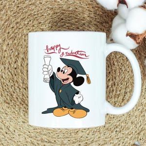 May include: White ceramic mug featuring Mickey Mouse in a graduation cap and gown, holding a diploma. The mug has the words "happy graduation" in red script. The handle is white.