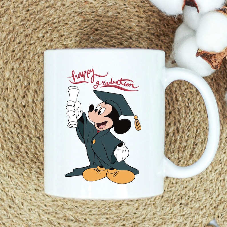 May include: White ceramic mug featuring Mickey Mouse in a graduation cap and gown, holding a diploma. The mug has the words "happy graduation" in red script. The handle is white.