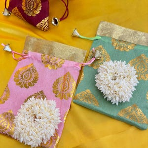 May include: A collection of small, decorative drawstring pouches in various colors, including pink, green, burgundy, and white. Each pouch features a gold paisley pattern and is adorned with a white floral embellishment. The pouches are arranged on a yellow fabric surface.