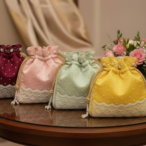 Peut inclure: Quatre sacs à cordon en satin bordeaux, rose, vert menthe et jaune. Chaque sac est orné d'une bordure perlée et d'une fermeture à cordon. Les sacs sont présentés sur une table ronde en bois.