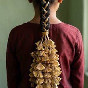 May include: A person with dark hair styled in a braid with gold accents and a decorative gold tassel. The person is wearing a maroon shirt, and the background is a muted green.