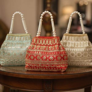 Puede incluir: Tres bolsos adornados con cuentas en azul pastel, rojo y crema, cada uno con un asa de perlas. Las bolsas están bordadas con hilo dorado y plateado, y están sobre una mesa redonda de madera. Las bolsas tienen un diseño tradicional y elegante.