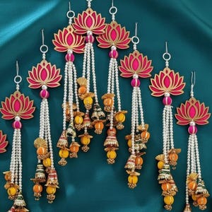 Könnte beinhalten: Dekorative Hängeornamente mit Lotusblüten-Designs in Rot und Gold. Jedes Ornament hat eine Schnur aus weißen Perlen mit bunten Quasten in Gelb, Orange und Grün. Der Hintergrund ist ein türkisfarbener Stoff.